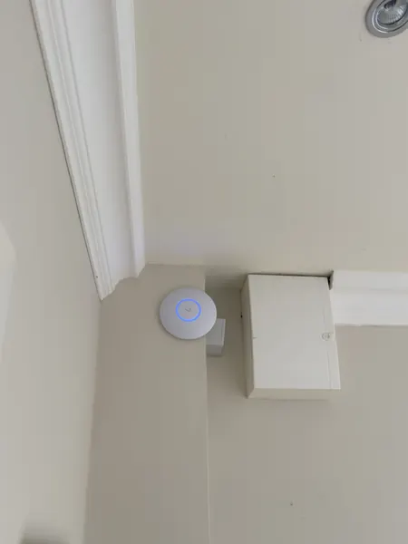 Access point with a blue light ring fixed high on a wall at ceiling level, next to a cream junction box