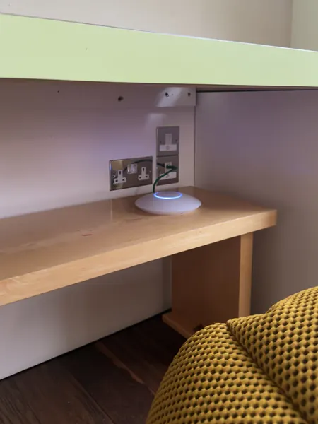 Round access point with a blue light ring sitting on a wooden shelf next to a socket, a green cable running to a data outlet