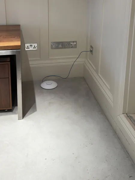 An old round access point left on the floor by a desk, a thin blue cable rising from it to a wall outlet