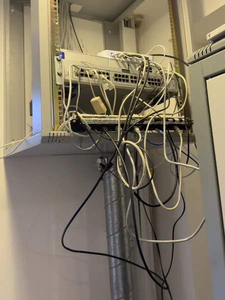 Network switches and a patch panel on a wall mounted frame, with white, black and blue cables hanging loose underneath