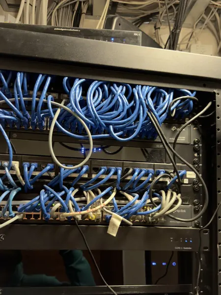 Overloaded rack seen from the front, its network switches buried under blue patch leads, power cables trailing down and a Control4 CORE 5 controller mounted beneath