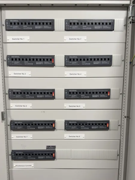 Row upon row of Dynalite relay units, numbered Switcher No.1 to No.8, fill an open cabinet above a miscellaneous controller