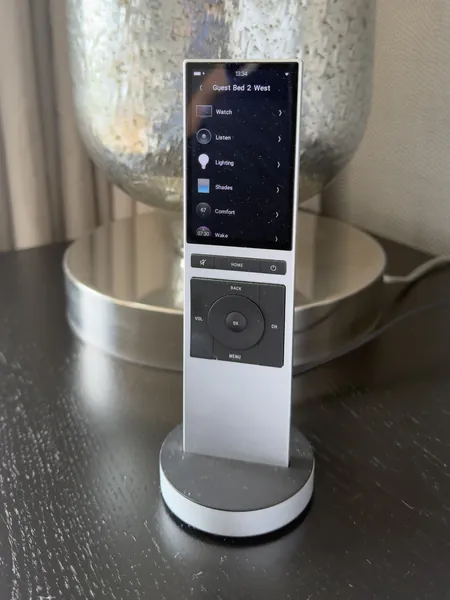 Guest bedroom remote standing in its charger by a silver glass lamp, its screen listing watch, listen, lighting, shades and comfort