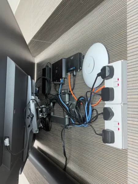 Crowded recess behind a television: sockets, a Wi-Fi access point, a Blustream unit, power supplies and coloured network cables