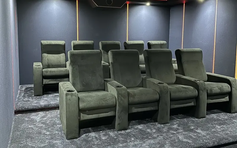 Two rows of green velvet recliners on a grey carpet in a dark home cinema, with amber light strips down the walls