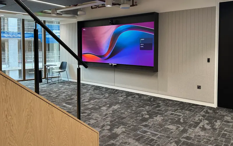 Large wall mounted display with a camera bar in an office meeting space, seen past a staircase handrail