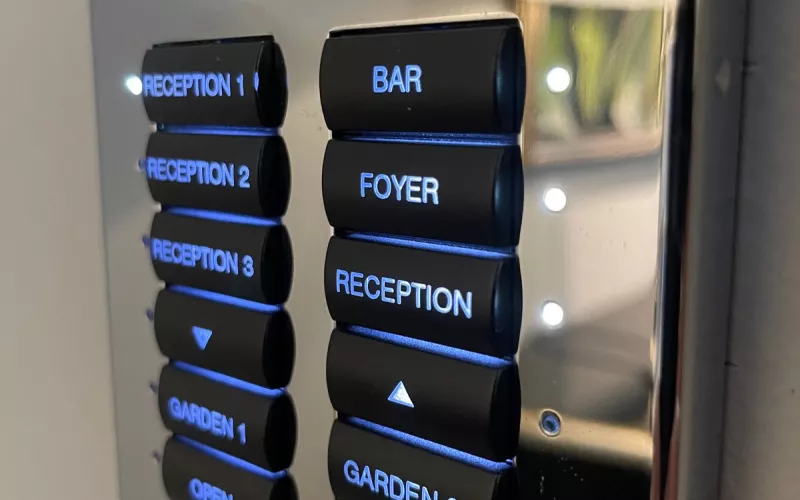 Lighting keypads in Surrey: a polished chrome keypad with black buttons engraved Reception 1, 2 and 3, Bar, Foyer, Garden and Open and Close, lit blue