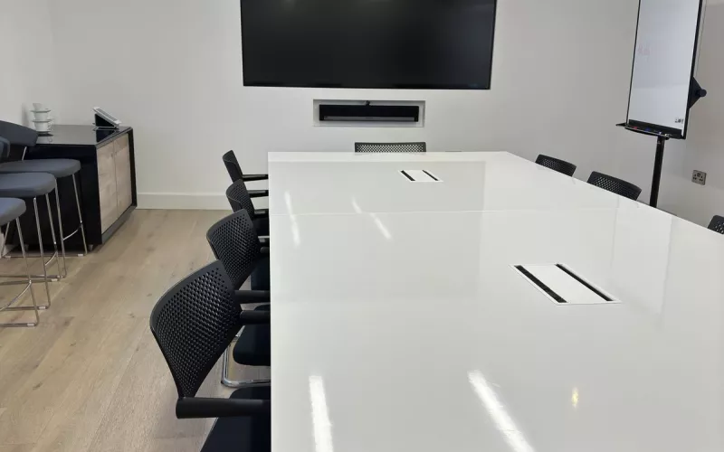 A meeting room wall display with a Sonos soundbar sunk into the wall beneath it and a small camera on top, facing a white table with black mesh chairs