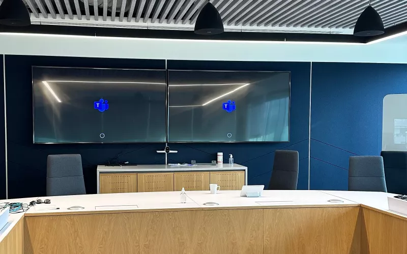 Boardroom AV installation: two displays side by side on a deep blue acoustic wall, the Teams logo on both and a camera on the credenza below