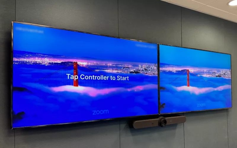 Zoom Rooms installation: twin screens on grey acoustic fabric, the Zoom logo on both and Tap Controller to Start on the left one, with the video bar underneath