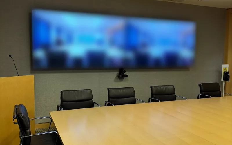 Boardroom video conferencing: a pair of screens on a grey wall, a camera beneath them and black leather chairs along a light timber table
