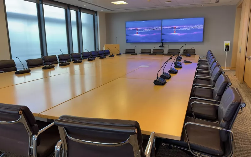 A boardroom audio system with microphone units running down both sides of a long timber table, and the Zoom Rooms start screen on the twin displays