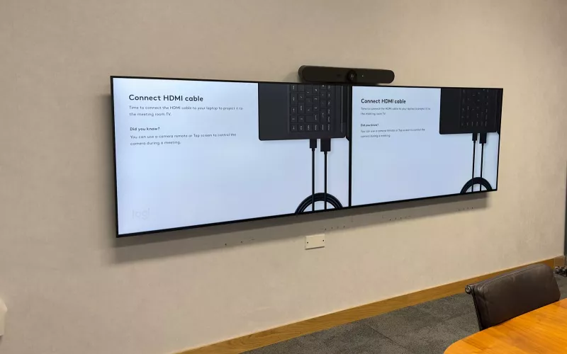 Meeting room technology on one wall: a Logitech video bar above two screens side by side, with the oval timber table in front