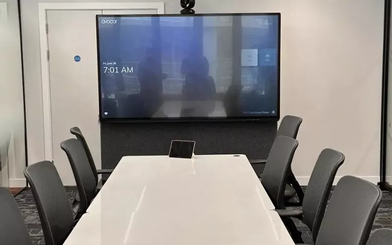 Glass walled meeting room with a wall mounted display, a camera above it, a long white table with a small touch panel and dark chairs, the kind of room where Teams Rooms Android devices are typically used