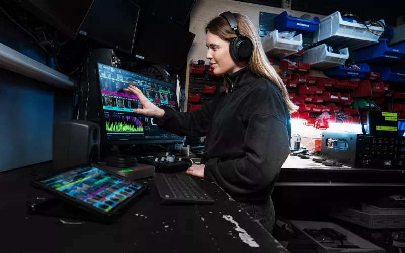Sound engineer in headphones backstage touching a monitor running Shure WAVETOOL 4, with an iPad on the desk and a wireless receiver on the bench