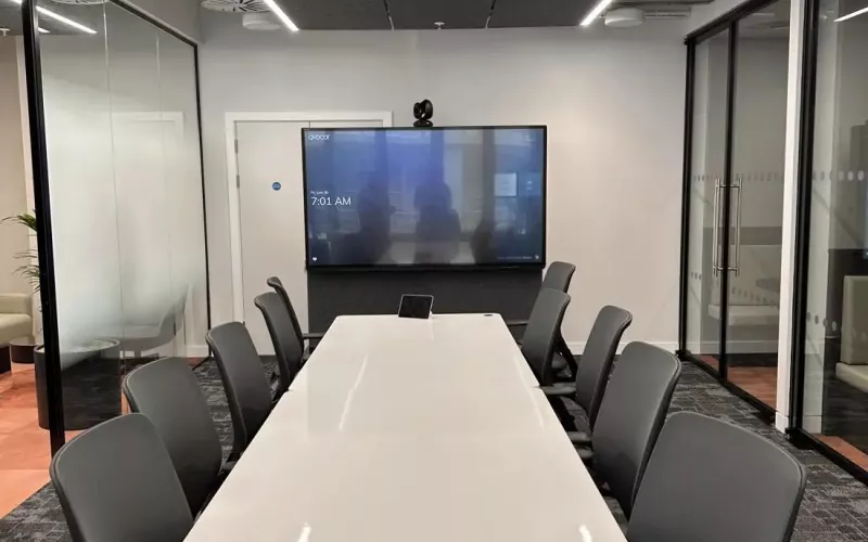 Glass walled meeting room with a wall mounted display and a camera above it, a long white table carrying a small touch panel, dark chairs and a ceiling of acoustic panels and linear lights, the kind of room where a Neat Teams certified controller would sit on the table