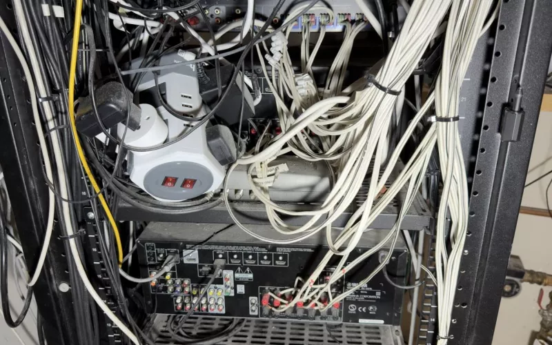 Control4 rescue, day one: behind a crowded rack, a stack of plug adaptors and a power strip sit over an AV receiver while thick white cable runs down the side to its speaker outputs