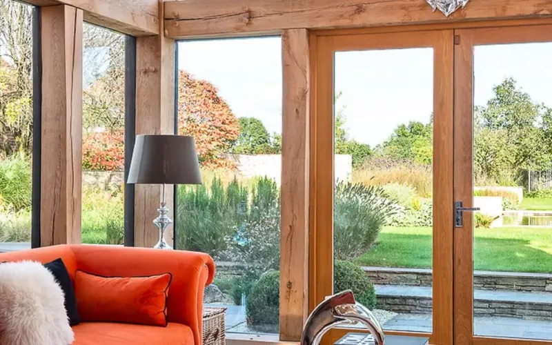 Oak framed garden room with tall glazing, glazed doors onto a landscaped garden and orange sofas, the kind of bright room where Crestron horizontal sheer shades soften daylight