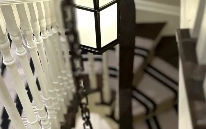 Lantern pendant hanging in a stairwell above a striped stair runner and white spindles, the kind of feature light a Lutron or Rako system dims