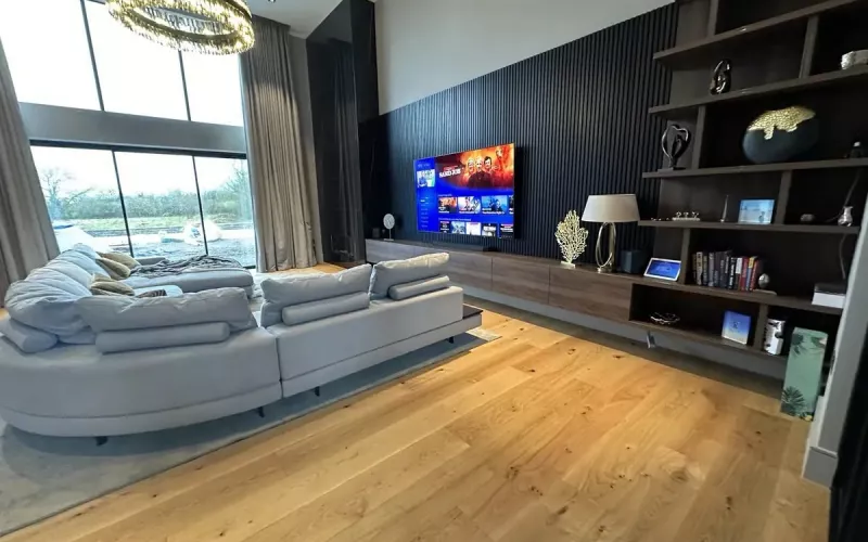 Double height living room with a curved grey sofa, an oak floor, a large TV on a dark slatted wall and tall windows, the kind of open space where ceiling speakers need careful spacing