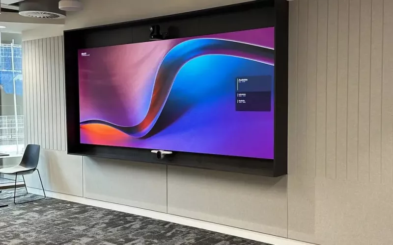 Wall screen in a dark surround with a camera on top in an open plan office beside tall windows, a typical position for interactive displays in an office