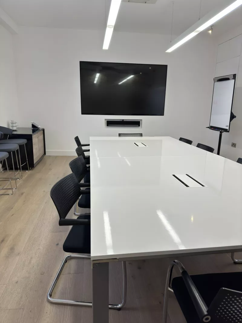 A meeting room wall display with a Sonos soundbar sunk into the wall beneath it and a small camera on top, facing a white table with black mesh chairs