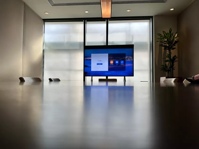 A Neat Board installation seen from down the table: the screen on its castor stand, the dark timber tabletop and daylight from the glazing behind
