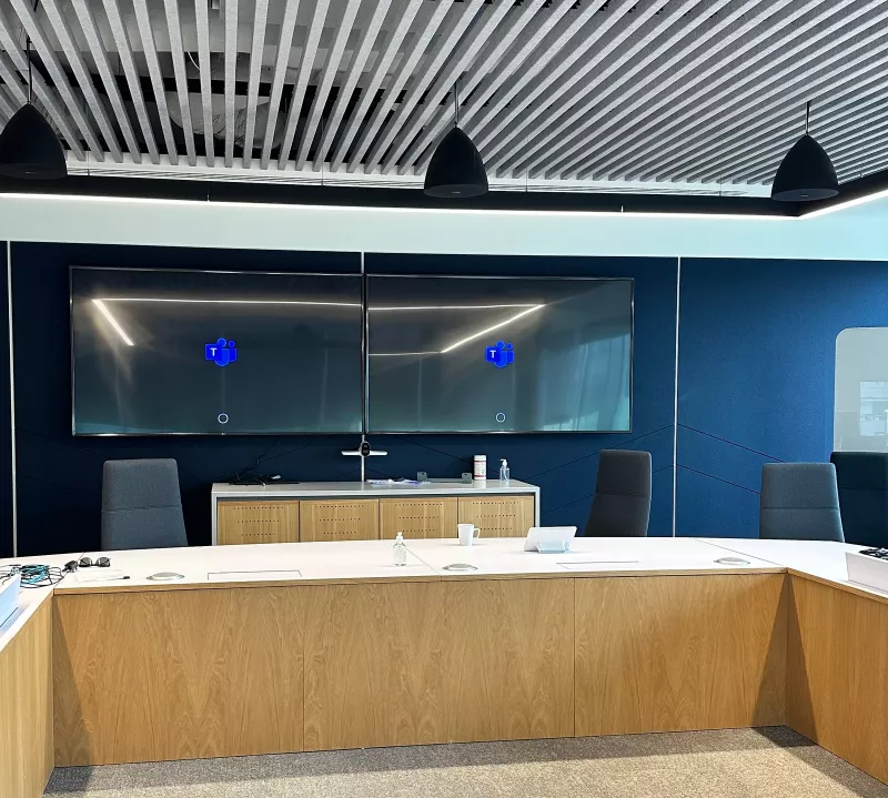 Boardroom AV installation: two displays side by side on a deep blue acoustic wall, the Teams logo on both and a camera on the credenza below