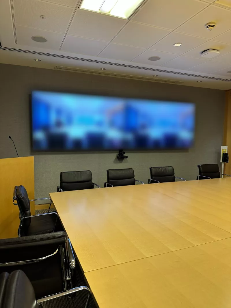Boardroom video conferencing: a pair of screens on a grey wall, a camera beneath them and black leather chairs along a light timber table