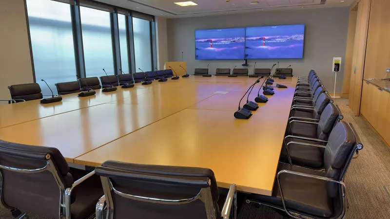 A boardroom audio system with microphone units running down both sides of a long timber table, and the Zoom Rooms start screen on the twin displays