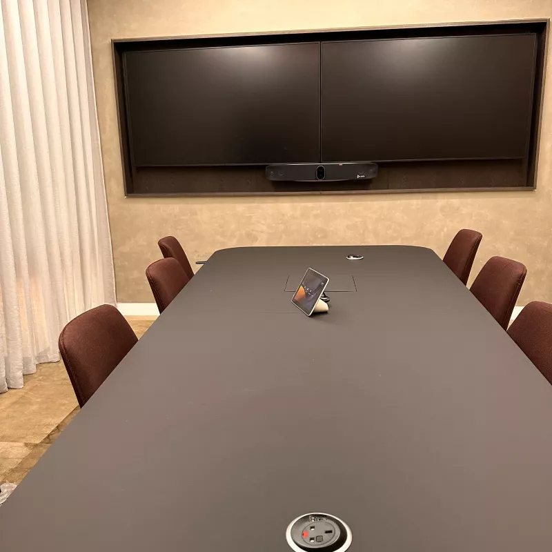 Long meeting table facing two wall displays and a Poly video bar, with a touch controller and a power socket let into the table