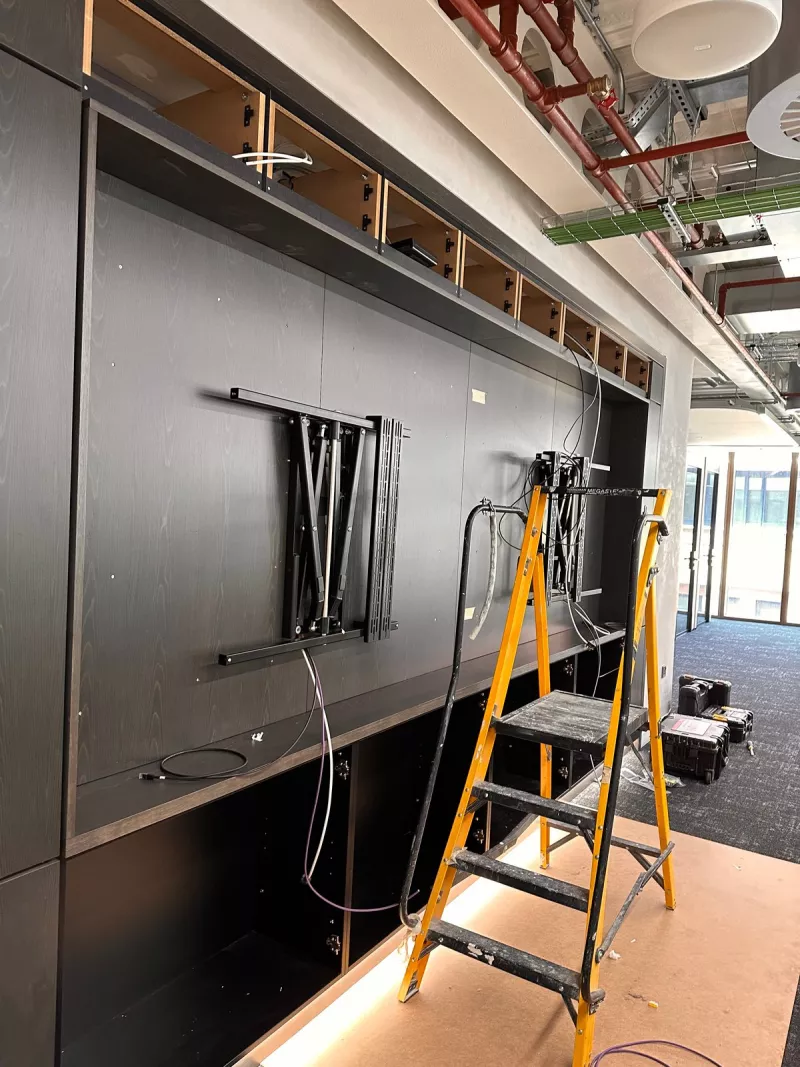 Two swing arm display mounts fixed to the back panel of a dark joinery wall in a London office, with network cables hanging beside them
