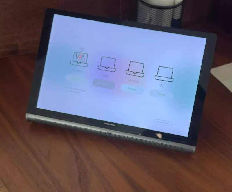 A Crestron touch panel with buttons for wireless Kramer VIA, table, lectern and PC sources, sitting on a dark wooden desk