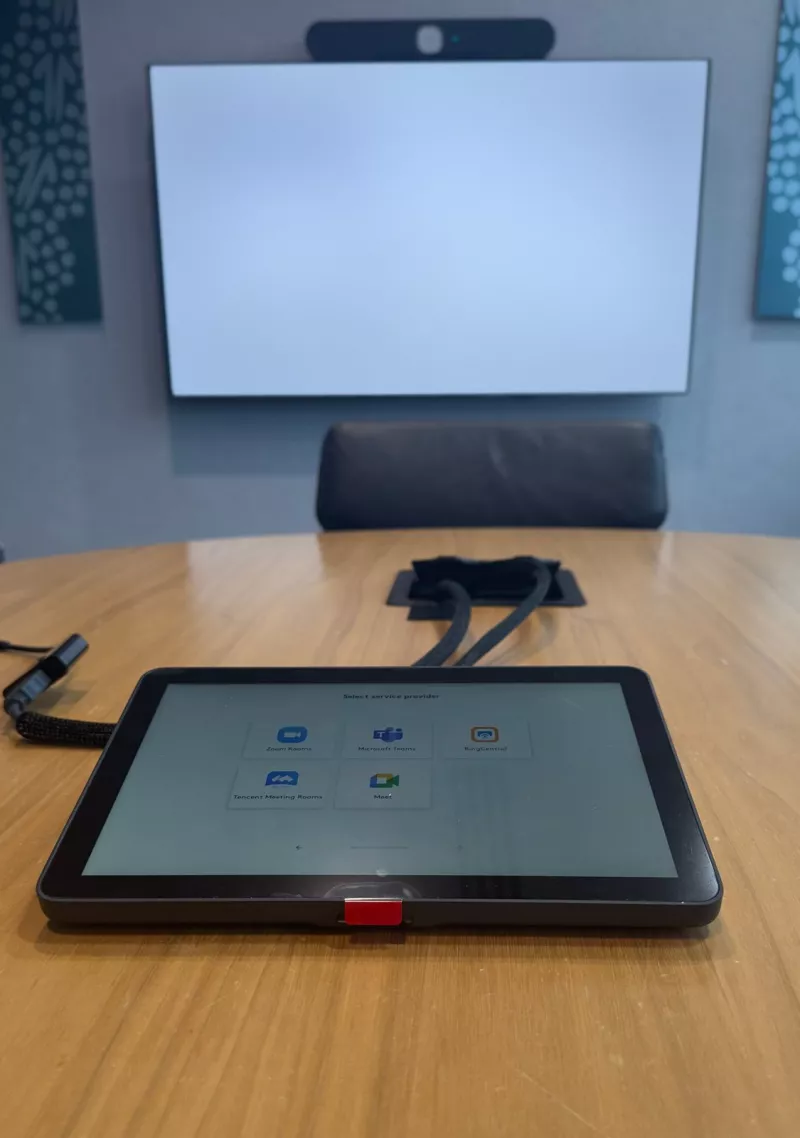 On a round timber table, a Logitech Tap showing its list of meeting services, with the room's screen and video bar out of focus behind