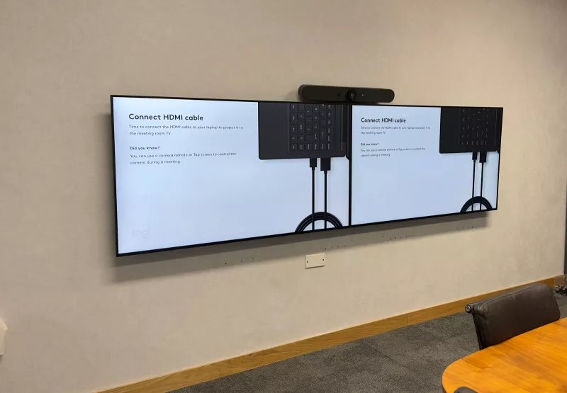 Meeting room technology on one wall: a Logitech video bar above two screens side by side, with the oval timber table in front