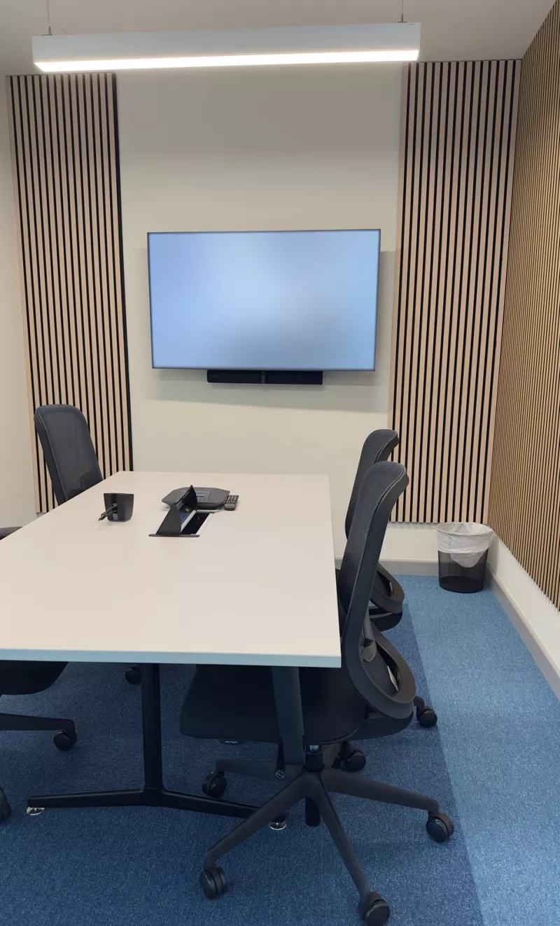 Slatted timber panels framing the display in a compact meeting room, with a slim bar under the screen and a phone in the middle of the white desk