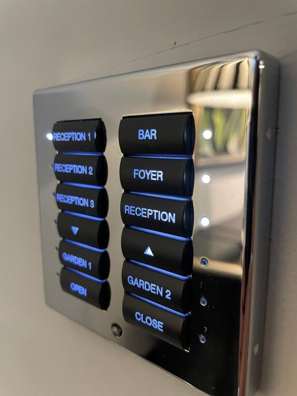 Lighting keypads in Surrey: a polished chrome keypad with black buttons engraved Reception 1, 2 and 3, Bar, Foyer, Garden and Open and Close, lit blue