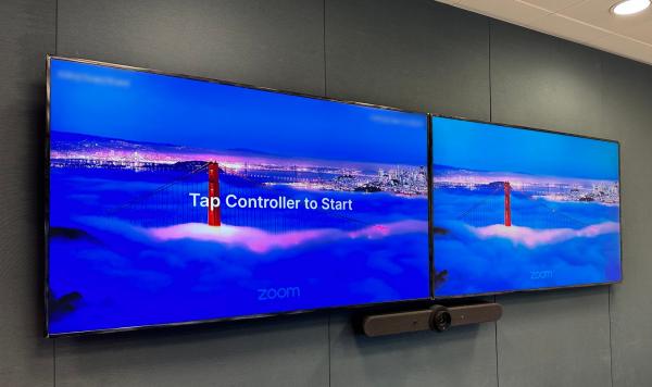 Zoom Rooms installation: twin screens on grey acoustic fabric, the Zoom logo on both and Tap Controller to Start on the left one, with the video bar underneath