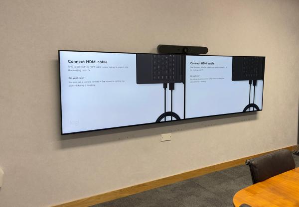 Meeting room technology on one wall: a Logitech video bar above two screens side by side, with the oval timber table in front