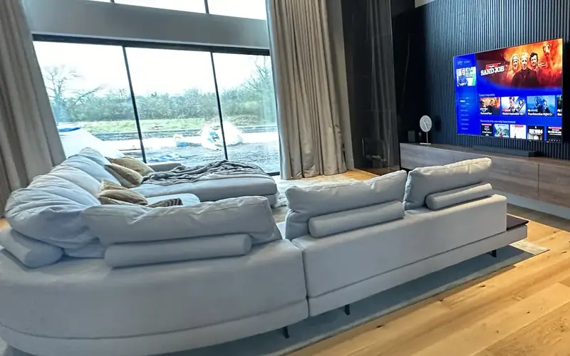 Large pale corner sofa in front of a tall window, with a TV on dark slatted panelling to one side